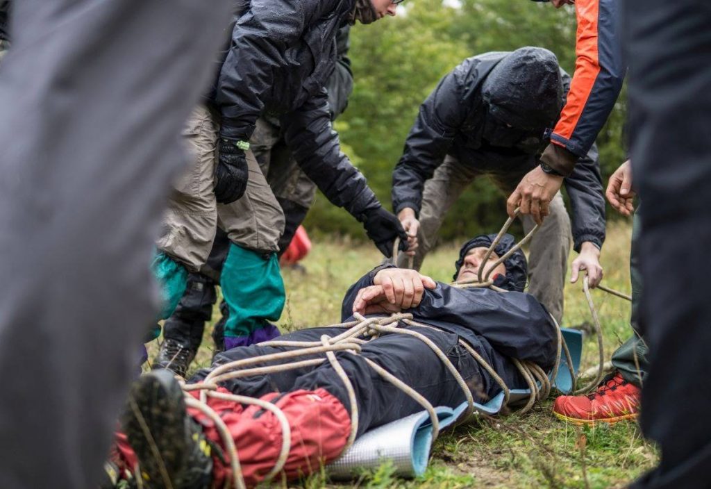 Zdjęcie mężczyzny leżącego na karimacie, spętanego sznurkami w formie noszy. Obok kilka osób w przeciwdeszczowych ubraniach. Zachpowują się jakby chcieli udzielić leżącemu pomocy/przygotować go do transportu