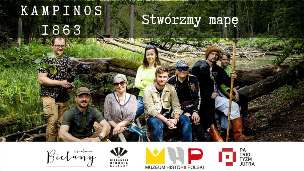 Zdjęcie zespołu realizującego projekt Kampinos 1863 na tle lasu (powalone drzewa i staw w pogodny letni dzień). 8 osób - 3 kobiety, 4 mężczyzn i chłopiec siedzą na przewróconym pniu drzewa i uśmiechają się do kamery. Jeden z mężczyzn przebrany jest w strój powstańca, trzyma w ręku kosę