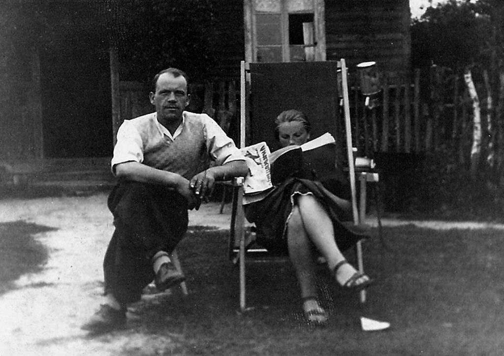 Czarnobiała fotografia mężczyzny i kobiety siedzących przed domem. Mężczyzna siedzi na stołku, opiera sie o leżak na którym siedzi kobieta.