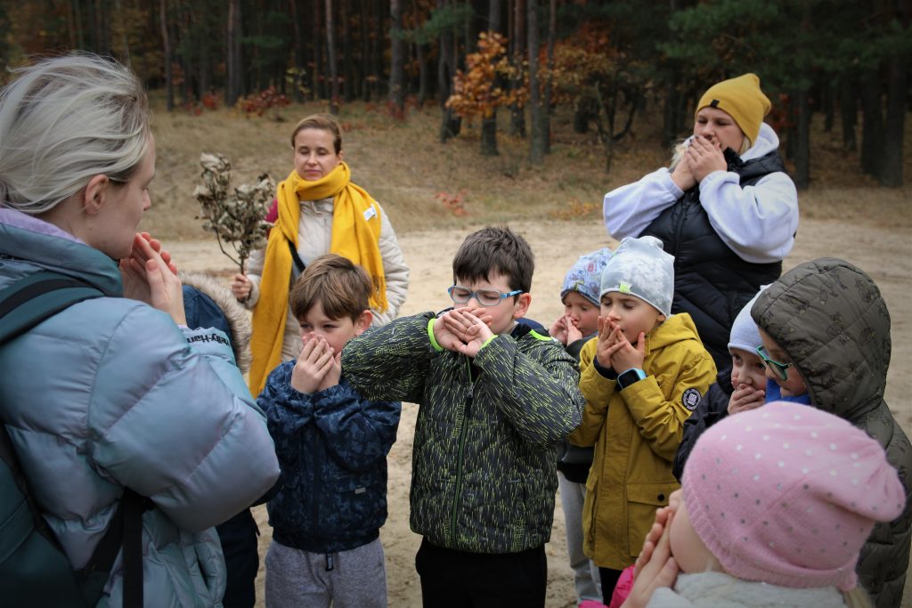 Grupa dzieci stojąca wraz z dorosłymi na piaszczystej polanie w lesie.. Zakrywają dwiema dłoniami usta równocześnie zamykając oczy.
