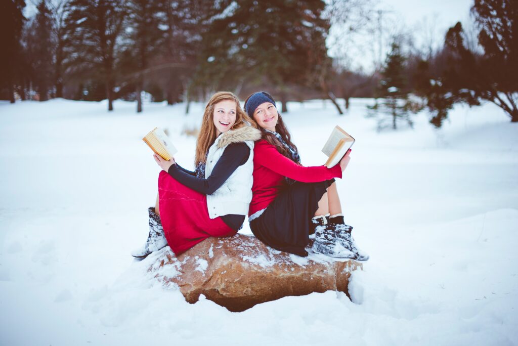 Dwie kobiety siedzą na kamieniu skierowane do siebie plecami. Obie trzymają książki. Cały kadr utrzymany jest w scenerii zimowej.