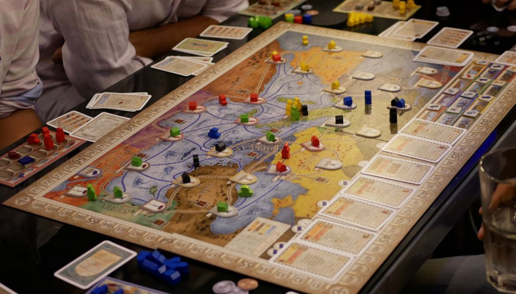 Na środku stołu leży rozłożona gra planszowa, której głównym elementem jest mapa świata śródziemnomorskiego. Ludzie siedzący dookoła blatu są uczestnikami gry.
