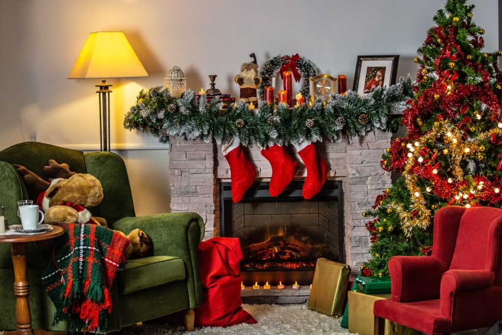 Pokój przyozdobiony świątecznie. W centrum fotografii znajduje się rozpalony kominek, na którym wiszą świąteczne ozdoby. Po bokach kadru stoja fotele - zielony oraz czerwony. Za czerwonym fotelem stoi ubrana wysoka choinka.