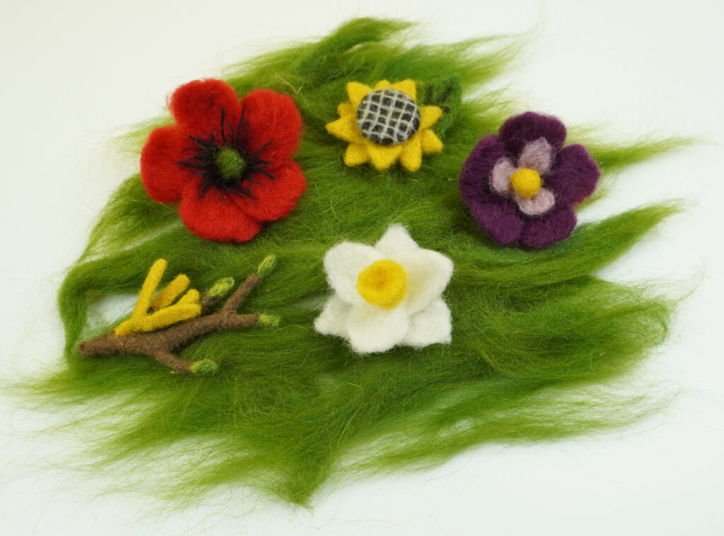 filcowe kwiaty - mak, słonecznik, bratek i kwiat o białych płatkach leżące wraz z filcową gałązką na filcowej, zielonej trawie