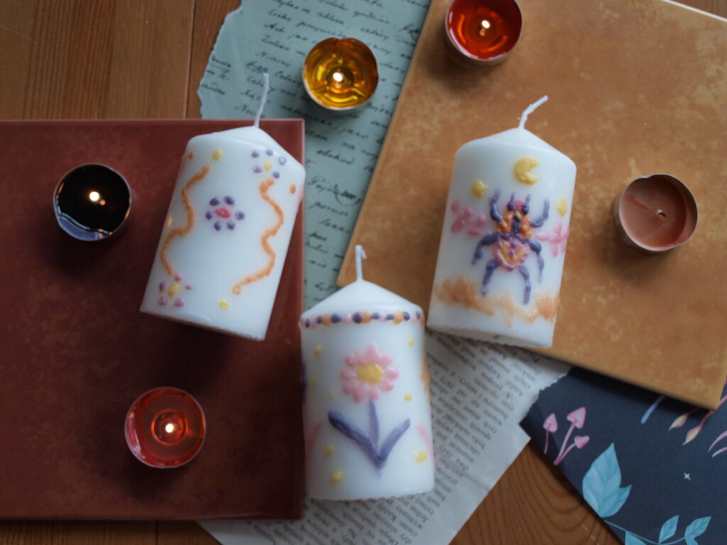 trzy białe świeczki pomalowane kolorowym woskiem - dwie w kwiatki, zygzaki i kropki i jedna z kolorowym żukiem