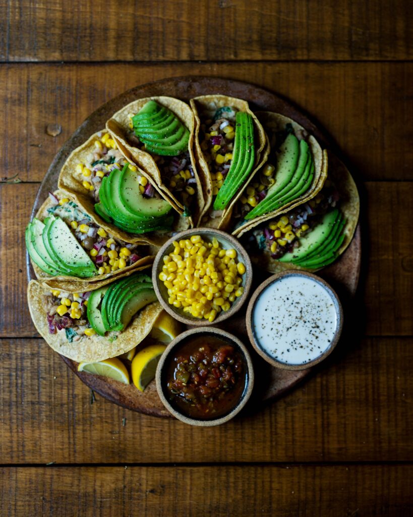 Leżący na drewnianym stole talerz pełen tortilli z avocado, kukurydzą i drobno posiekanymi warzywami. Obok tortilli na talerzu znajdują się miseczki z kukurydzą i sosami do wrapów.