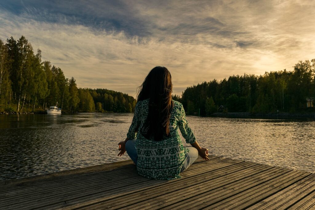medytująca kobieta siedząca po turecku, plecami do obiektywu. Kobieta siedzi na drewnianym pomoście nad jeziorem. W tle widać zielone drzewa i błękitne, lekko zachmurzone niebo.